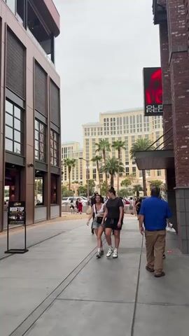 The video shows a sidewalk scene with several people walking. On the left, there is a modern building with large windows. On the right, there is a brick building with a sign that says "OLD RED." In the background, there are tall buildings and palm trees. The sky is overcast. Two women are walking in the foreground, and a man in a blue shirt is walking away from the camera. Other people are visible further down the sidewalk. There are also vehicles and fountains in the background.