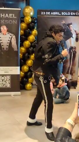The video opens with a static shot of a Michael Jackson impersonator standing center frame, facing away from the camera. The impersonator is dressed in a black jacket, white shirt, black pants with a stripe down the side, white socks, and black shoes. Behind him are two large banners, one on the left featuring Michael Jackson's image and the text "MICHAEL JACKSON," and another on the right with text that appears to be an event announcement. A cluster of black and gold balloons is situated between the banners. To the right of the impersonator, a man in a black jacket and jeans is standing, looking towards the impersonator. In front of the man, two young children are seated on the floor, a girl in a pink outfit and a girl in a denim jacket. The impersonator then turns to face the camera and begins to sing or speak into a microphone he is holding. The camera then zooms in slightly and pans to the right, following the impersonator's movements as he performs. He gestures with his free hand and moves his body. The background remains largely the same, with the banners, balloons, and other people visible. The video ends with the impersonator in mid-performance.