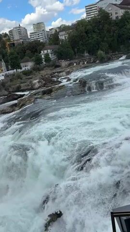 A wide shot captures the powerful Rhine Falls in Switzerland. The water churns and crashes over rocks, creating a mist that rises into the air. In the background, buildings and trees line the riverbank, with a red train visible on a track. Several boats are present on the water, including a yellow tour boat near the falls. The sky is partly cloudy with a blue sky peeking through. The video pans slightly, offering a dynamic view of the natural wonder. No people are clearly visible in the foreground, but there appear to be some individuals on a viewing platform or walkway in the distance. The overall scene is one of natural power and beauty, with human elements integrated into the landscape.