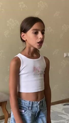 A young girl stands in front of a floral wallpapered wall, wearing a white crop top with a cartoon bear graphic and distressed denim shorts. She is looking off to her right, with her mouth slightly open as if speaking or singing. Her hair is pulled back in a ponytail. The room appears to be indoors, with a light-colored floor and what looks like a stool to the left of the frame. The lighting suggests it is daytime. The graphic on her shirt has the word "BESTDAY" written on it.