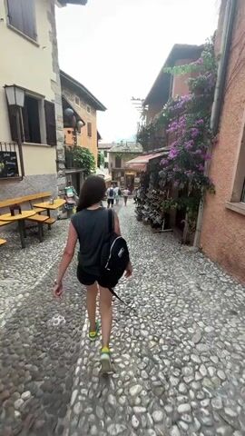 A young person with long dark hair, wearing a dark grey tank top, black shorts, and neon green and yellow sneakers, walks away from the camera down a cobblestone street. They are carrying a black backpack. The street is lined with buildings on both sides. On the left, there are tables and benches outside a building with wooden shutters. On the right, there are shops with awnings, displaying merchandise such as shoes and bags. Purple flowers cascade down the building on the right. Other people are visible further down the street, walking in the same direction. The sky is overcast.