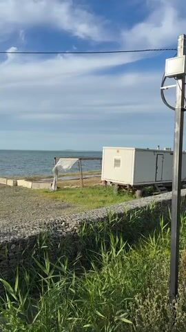 The video presents a panoramic view of a coastal area in Taman, Russia, under a partly cloudy blue sky.

Initially, the shot frames a white mobile container or trailer situated near the water's edge, alongside a makeshift wooden shelter covered with white fabric. The foreground features a gravelly ground, a gabion wall filled with stones, and green reeds.

As the camera slowly pans to the left, the white container moves out of view. A tall pole equipped with multiple CCTV cameras and a red box becomes visible on the left side of the frame. In the immediate foreground, a paved walkway, partially made of wood, and a black metal railing are seen. The pan concludes with a wide shot of the calm body of water stretching to the horizon, under a bright, daytime sky. No people or specific activities are visible in the scene.