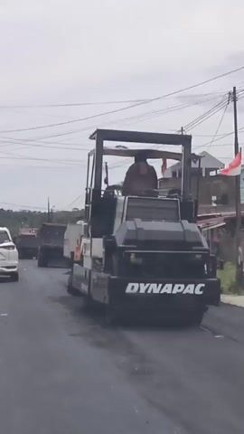 A roller machine with the brand name DYNAPAC is seen paving a road. The machine is black and white and has a driver in the cabin. Several dump trucks are visible behind the roller machine, presumably delivering asphalt. A white SUV is seen in the background, driving in the opposite direction. The sky is overcast. The scene appears to be an ongoing road construction or repair project.