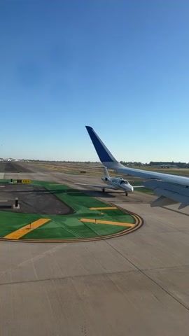 The video shows an aerial view of an airport tarmac. In the foreground, the wing of a large passenger airplane is visible, angled towards the right side of the frame. In the middle ground, a smaller, white private jet is positioned on the tarmac, facing the viewer. The private jet is on a taxiway marked with green and yellow lines. In the background, a runway stretches into the distance, with more taxiways and some distant buildings. The sky is clear and blue. The camera appears to be mounted on the passenger airplane, looking out its window.
