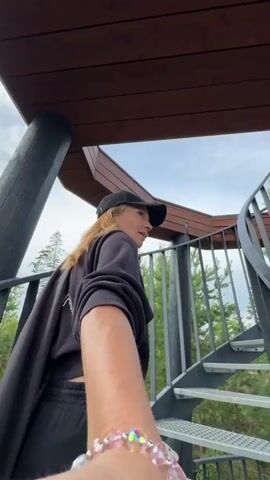 A person wearing a black baseball cap and a black sweatshirt with a towel draped over their shoulders walks up a spiral staircase. The staircase is made of metal with a wooden railing and is outdoors, surrounded by trees. The sky is cloudy. The person is captured from the perspective of someone walking ahead of them and holding their hand.