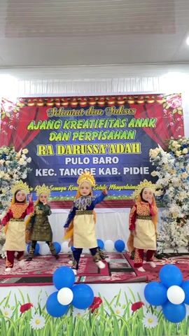 This media file captures a dance performance by young children on a stage, likely at an event in Keudetangse, Indonesia. Five children, dressed in elaborate traditional attire with gold crowns, are performing a synchronized dance. Their costumes consist of richly embroidered tunics and skirts, with some wearing blue and others red tops. The stage is decorated with a large banner that reads "SELAMAT dan SUKSES AJANG KREATIFITAS ANAK DAN PERPISAHAN RA DARUSSA' ADAH PULO BARO KEC. TANGSE KAB. PIDIE." Below this, there is smaller text.

The backdrop behind the performers features a red and gold floral design. Flanking the stage are tall, white floral arrangements that add to the festive atmosphere. The stage floor is covered with a red carpet, and blue and white balloons are scattered around, with clusters of balloons in blue and white also placed at the front of the stage and on the floor. The lighting suggests an indoor setting, possibly during the daytime. The children appear focused and engaged in their performance, embodying a sense of youthful energy and cultural pride. The overall scene is one of celebration and a showcase of young talent.