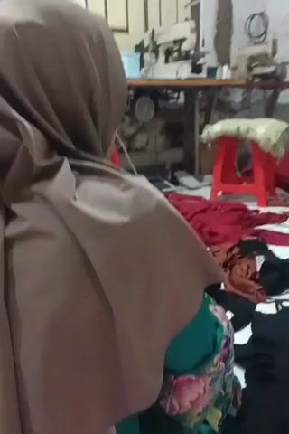 The video shows a woman wearing a brown hijab. She is sitting down and appears to be holding a baby. The background is blurry but seems to be an indoor space with some sewing equipment and fabric. The woman's hands are visible, and she is gently touching the baby. Another person is partially visible in the background, also wearing a hijab. The lighting is dim.