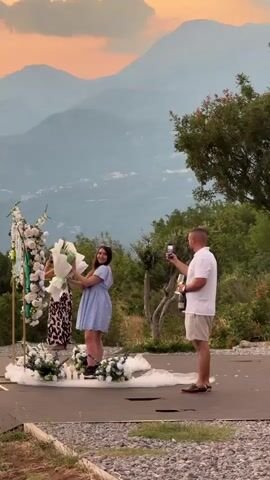 This media file captures a romantic proposal with a breathtaking mountain backdrop at sunset in Čelobrdo, Montenegro. Three individuals are present: a woman in a light blue dress holding a bouquet of white and green flowers, a person behind her in a leopard print dress, and a man standing to the right. The man is holding a smartphone, seemingly recording the moment, and has a bottle of champagne in his other hand, suggesting a celebration.

The scene is set on an elevated platform, possibly a viewpoint or an outdoor event space, overlooking rolling mountains bathed in the warm, soft light of sunset. The sky displays hues of orange and pink, characteristic of the golden hour. In the foreground, a decorative setup for a proposal is visible, featuring a golden arch adorned with white and green flowers, white tulle fabric, and lit candles scattered around, creating an intimate and celebratory atmosphere. Lush greenery and trees frame the right side of the scene, adding to the natural beauty of the location.

The woman in the blue dress appears to be the focus of the proposal, holding her bouquet with a joyful expression. Her posture and gaze suggest she is interacting with the man holding the phone. The overall mood is joyful, romantic, and celebratory. The presence of the champagne bottle and the recording device indicates a significant event, most likely an engagement proposal. No visible text is present in the media file.