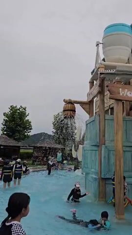 A large water bucket at a water park is tipping over, spilling a torrent of water into a pool below. Several children and adults are in the pool, some playing and others reacting to the falling water. The water bucket structure is wooden and weathered, with a sign that reads "Water Bomb." Green wooden crates are stacked around the base of the structure. The sky is overcast.