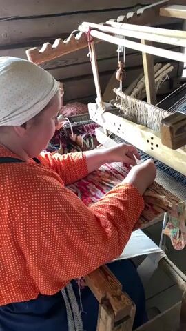 A woman wearing a white headscarf and an orange polka-dot blouse is seated at a wooden loom, actively weaving. Her hands are engaged with the threads, guiding them through the intricate process. The loom itself is a complex wooden structure with numerous threads stretched vertically and horizontally. A partially woven fabric, displaying a colorful pattern of red, pink, and yellow stripes, is visible on the loom. To the woman's right, a pair of scissors rests on the woven material. The background consists of wooden planks, suggesting an indoor setting, possibly a workshop or a traditional craft space.