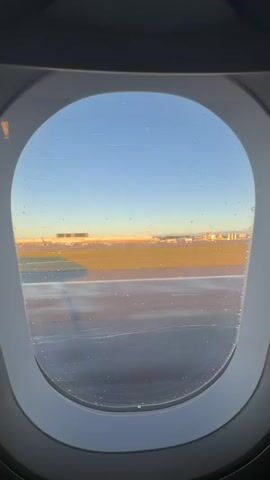 The video captures a serene view from an airplane window, looking out onto an airport scene during what appears to be the golden hour or early morning/late afternoon. The oval-shaped window frame dominates the foreground, with numerous small water droplets or smudges visible on the glass.

Outside the window, a wide expanse of asphalt runway or taxiway stretches horizontally across the lower part of the frame, showing movement past the window, suggesting the aircraft is taxiing. Beyond the paved area, a strip of golden-brown grassy land separates the foreground from the distant airport infrastructure. In the background, several buildings and what looks like an airport control tower are silhouetted against a clear, light blue sky. Another aircraft can be briefly seen in the distance as the plane moves. The warm, low-angle sunlight casts a soft glow over the landscape, highlighting the textures of the ground and the distant structures. The overall impression is one of calm and preparation for travel. The audio features the distinct hum of an airplane engine.