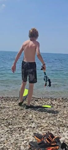 A shirtless young person stands on a rocky beach, facing the ocean. They are wearing patterned shorts and black fins on their feet. In their right hand, they hold a snorkel mask and snorkel. The ocean is blue and calm under a clear sky. The person takes a step towards the water, with one fin already in the shallows. Another fin is visible on the ground behind them.