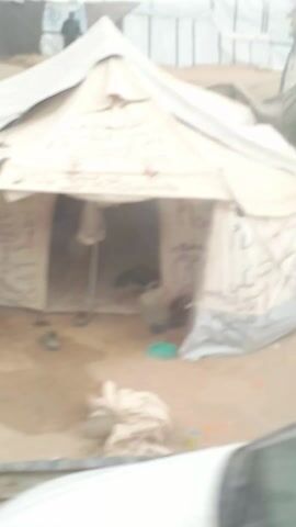 The video depicts a refugee camp with numerous tents, primarily white or light-colored. The tents appear worn and some are marked with Arabic writing. The ground is sandy and littered with debris. There are a few people visible, including children, moving within the camp. The video is taken from a moving vehicle. A piece of paper passes in front of the camera briefly.