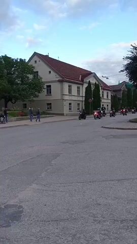 A group of motorcyclists ride down a street in front of a building. The motorcyclists are riding in formation, and the motorcycles are all facing the same direction. The building is a large, two-story building with a red roof. There are trees in front of the building, and there are people walking on the sidewalk in front of the building. The sky is cloudy.