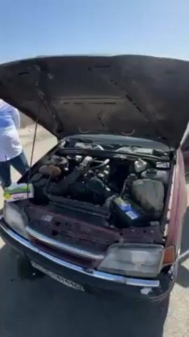 The video shows a maroon sedan with its hood open, parked on the side of a desert road on a sunny day. A person wearing a white shirt and jeans is standing near the car. The road stretches straight into the distance, shimmering in the heat. The car's driver-side door is open, revealing the interior. The environment is arid and sparsely vegetated. The car appears to be undergoing maintenance or experiencing mechanical issues.