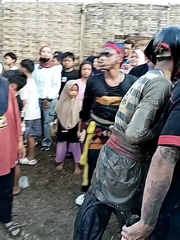 The video shows a muddy spectacle taking place in what appears to be an enclosed area with a bamboo fence. Several people are gathered as spectators, including adults and children. Some individuals are wearing face masks. A group of men covered in mud are the apparent focus of the event, engaging in some sort of activity within the muddy enclosure. One man stands off to the side, seemingly overseeing the proceedings. The sky is visible in the background, with some clouds.