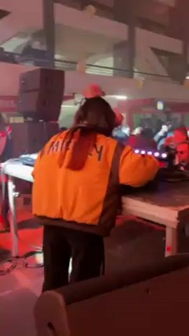 The video captures the atmosphere of a lively event, possibly a concert or party, with music playing in the background. The primary focus is on two individuals: a DJ and a man in the crowd. The DJ, wearing a distinctive yellow and black jacket with "Hustle" written on the back, stands behind a DJ booth, likely mixing music. In the crowd, a man wearing sunglasses, a cap, and a black puffer jacket raises his middle finger while holding a drink. The environment is a large, open space with visible beams and overhead structures, suggesting a warehouse-type venue. The crowd is dense and energetic, with people moving and enjoying the music. Red lighting and smoke effects enhance the party atmosphere.