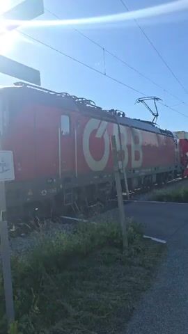 A train is moving from left to right. The train is red and yellow and has a logo with the letters "BB" on it. The train is on a track that crosses a road. There is a red and white crossbuck sign next to the track. The sky is blue and there are some clouds. The sun is shining. There is grass on either side of the track.