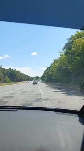 The video shows a point-of-view shot from inside a vehicle driving on a highway. The road is paved and has lane markings. There are several other vehicles on the road, including cars and a white van. The landscape on either side of the road is lined with trees. The sky is blue with a few scattered clouds, indicating fair weather. The video appears to be taken during the daytime.