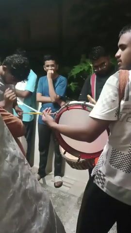 The video shows a group of young men playing drums outdoors at night. The foreground is dominated by a young man playing a large drum with drumsticks. He is wearing a white t-shirt with a checkered pattern. Behind him, several other young men are visible. One is playing a smaller drum, another is holding drumsticks, and a third is looking on. The environment appears to be a paved area with some foliage in the background. The lighting is dim, suggesting it is evening or night. The men appear to be engaged in a musical performance or practice.