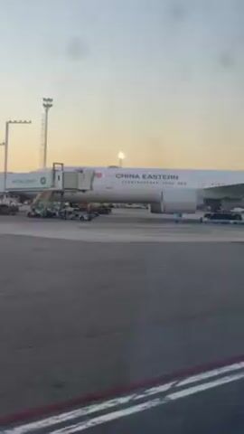 A passenger's perspective from inside a vehicle provides a view of a China Eastern airplane parked at an airport terminal. The aircraft is white with "CHINA EASTERN" printed along the fuselage. A jet bridge, marked "INTERCARGO," is connected to the plane. Various airport vehicles and ground staff are visible around the plane. The sky is clear and light, suggesting it might be early morning or late afternoon. The overall setting is a busy airport environment.