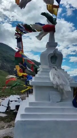 A white stupa is decorated with prayer flags. The stupa is on a rocky terrain with mountains in the background. The sky is blue with white clouds. The prayer flags are multi-colored and flutter in the wind. The stupa has a layered base and a domed top. There are white cloths wrapped around the stupa. The scene is outdoors during the day.