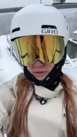 A person on a ski lift is filming a selfie video in snowy conditions. The individual wears a white Giro helmet, gold mirrored goggles, a black neck gaiter, and a light-colored ski jacket with visible snow accumulation. Long red hair is visible. The background shows snowy trees, other ski lifts, and a building partially obscured by snow. Snow is falling as the ski lift ascends.
