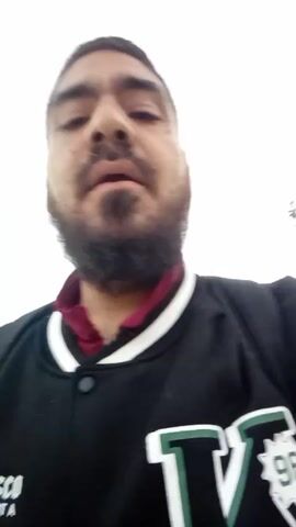 The video is a low-angle shot of a man with a dark beard. He is wearing a black jacket with green and white accents over a maroon collared shirt. He is looking directly at the camera and speaking. The background is a blurry white sky.
