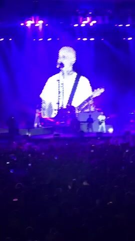 A large concert screen displays a pixelated image of a bald man wearing a white t-shirt and playing a guitar. Below the screen, on stage, three musicians are visible, two of whom appear to be playing instruments. The stage is lit with bright purple lights. In the foreground, a large crowd of people stands in darkness, many holding up their phones. The overall atmosphere is of a live music performance.
