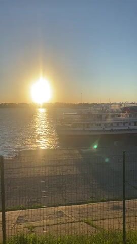 The video captures a sunset scene on a body of water, likely a river, in Novokuybyshevsk, Russia. The sun, a bright, intense orb, hangs low in the sky, casting a warm, golden glow across the rippling water. The light reflects off the surface, creating a shimmering path.

Dominating the middle ground of the frame is a large, multi-deck passenger ship, docked near the shore. The ship is white with red accents and appears to be a river cruise liner. Its numerous windows suggest it has significant passenger capacity. Lifeboats are visible on its upper decks.

In the foreground, a chain-link fence runs horizontally, separating the viewer from the dock area. Beyond the fence, a concrete embankment slopes down to the water. The ground appears to be a paved area with some scattered debris. Patches of green grass and foliage are visible at the very bottom of the frame, suggesting the presence of vegetation.

The sky is a clear, brilliant blue, transitioning to a lighter hue near the horizon where the sun is setting. The overall atmosphere is peaceful and serene, evoking a sense of calm as the day concludes. The time of day is clearly sunset, indicated by the position and intensity of the sun. There is no visible text in the video.