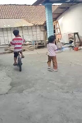 Two children are in a dirt yard. On the left, a young boy wearing a red and white striped shirt and white shorts is riding a bicycle. On the right, a young girl wearing a pink t-shirt and brown pants stands looking towards the boy. Behind them are several buildings with tile roofs and corrugated metal walls.
