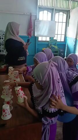 A group of young girls wearing purple hijabs are engaged in a cup-stacking activity. They are gathered around a wooden table, where several stacks of white paper cups decorated with red polka dots are arranged. A woman, possibly a teacher, stands to the left, holding a phone and addressing the girls. The girls are actively participating, with some reaching for cups, others holding cups, and one appearing to be in the process of adding a cup to a stack. The environment appears to be a classroom or similar educational setting, with a whiteboard and windows visible in the background. The atmosphere seems to be one of focused activity and learning.