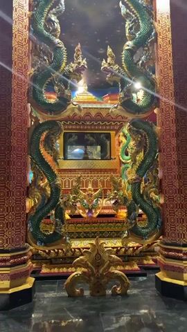 The video shows a view of an ornate structure, likely a temple or shrine, with intricate gold and green dragon carvings flanking a central altar. The altar itself is multi-tiered, adorned with smaller golden sculptures and a mirrored compartment at its center. The ceiling is dark and appears to be decorated with a starry pattern, with a golden pyramid-like structure visible at the apex. The lighting is dim, highlighting the metallic sheen of the carvings and decorations.