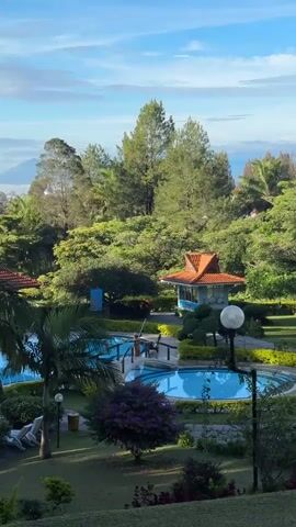 The video shows an aerial view of a resort or hotel with multiple swimming pools. The pools are blue and surrounded by lush green vegetation, including palm trees and manicured gardens. In the background, there are tall trees, a small building with an orange roof, and mountains in the distance. The sky is blue with some clouds. The lighting suggests it is daytime, likely morning or late afternoon. There are no people visible in the video, and no actions or interactions are taking place.