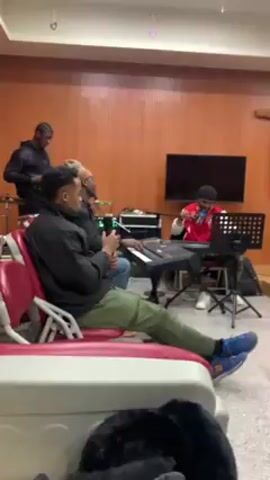 The video shows a group of young people, predominantly Black, in what appears to be a music rehearsal or worship setting. In the initial shot, some individuals are seated in red chairs, while others are standing or seated near musical equipment. There is a keyboard player in a red jacket, and a person with a microphone in front of them. A cross can be seen on the wall. The camera pans to the right, showing more people seated. One person holds what looks like a smartphone. A guitar stands next to a table, and a person stands near what might be a drum set. The general impression is that they are preparing to play or sing together. The environment appears to be a room with wood-paneled walls and tiled floors.