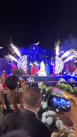 The video shows a group of women dancing on a stage, likely at a wedding celebration. The central figure wears a red and white lehenga, a traditional Indian wedding dress, while the other women wear matching red outfits. They hold small white objects in their hands, possibly for the dance. The stage is decorated with lights, a crescent moon, and a statue of a bull, likely Nandi, the mount of the Hindu god Shiva. Sparklers erupt on either side of the stage. People in the audience record the performance with their phones. The event appears to be taking place at night.