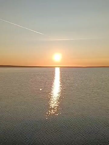 A wide shot of a calm body of water under a clear sky at sunset. The sun, a bright orange orb, is low on the horizon, casting a shimmering path of light across the water. The sky is a gradient of pale blue at the top, transitioning to soft orange and yellow near the horizon. A faint white contrail from an aircraft stretches diagonally across the upper left quadrant of the sky. The water is calm with gentle ripples reflecting the sunlight. The horizon line is flat and indistinct, suggesting a large, open expanse of water meeting the land or sky. No people or objects are visible in or on the water. The overall impression is one of tranquility and natural beauty.