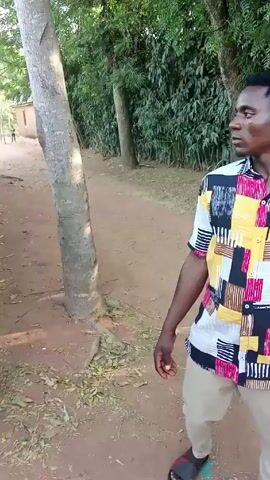 The video begins with a man walking on a dirt path, alongside trees. He wears a multi-colored shirt and light-colored pants.  The camera then moves to a woman with brown hair, wearing a blue t-shirt with "CossFit '04" printed on the back, and jeans. She is standing near a tree and a metal gate.  The woman turns, smiles, and gestures with her hands while facing the camera. She is standing on a dirt path, surrounded by trees and a building visible in the background.