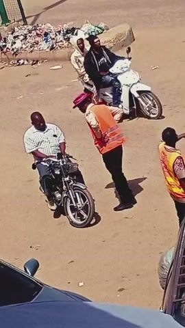 At timestamp 00:00, the scene shows a man seated on a motorcycle, facing left. A person wearing an orange safety vest and a pink hat is leaning towards the man on the motorcycle, appearing to speak to him. In the background, there is a pile of trash and a curb. At timestamp 00:01, the person in the orange vest extends their hand towards the man on the motorcycle. At timestamp 00:02, another person, wearing a black shirt and a green crate on their head filled with bottles, walks past in the background. The interaction between the man on the motorcycle and the person in the vest continues. At timestamp 00:03, the person with the crate of bottles turns and holds the crate up. Another person in a black t-shirt and black pants stands to the left of the motorcycle. At timestamp 00:04, the person in the orange vest is now holding a stick and is gesturing with it. The man on the motorcycle remains seated, and the person in the black t-shirt is still standing to the left.