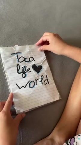 The video shows a person's hands interacting with a handmade paper dollhouse. The dollhouse is made from lined paper and laminated. The cover of the dollhouse has "Toca life world" written on it with a heart. The dollhouse opens to reveal two pages. The left page depicts a pink bed with a Hello Kitty design, a closet, and some accessories. The right page shows a wooden closet with shoes lined up at the bottom, and a hanger with a pink bunny. The person is seen opening the closet and placing a paper doll into it. The doll is wearing a brown dress and holding a teddy bear. The background is a gray textured surface, possibly a sofa or carpet.