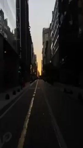 The video presents a ground-level view of a city street, apparently at either dusk or dawn, given the low-angle light. The street is flanked by tall buildings, casting much of the scene into shadow, although the buildings are visible in silhouette. The pavement is patterned, consisting of small bricks or similar pavers, and a painted white stripe runs along the right side, marking a lane. There are traffic cones and bollards on either side of the street. A sign for "Ministerio de Justicia" is visible on the right. At the far end of the street, a bright light source is visible, indicating the sun rising or setting. A few pedestrians are faintly visible, mostly standing or moving slowly. The overall impression is one of a quiet, possibly early morning, urban scene.