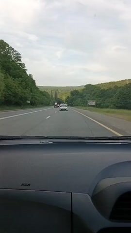 The video is a first-person perspective from inside a moving car, looking through the windshield. The car is on a highway with two lanes going in the same direction, separated by a dashed white line, and a solid yellow line on the right shoulder. Trees line both sides of the road, forming a green canopy. The sky is partly cloudy. Several other vehicles are visible on the road ahead, including a white sedan directly in front, a white truck in the left lane, and a black sedan on the far left. The dashboard of the car is visible in the foreground, with the "SRS airbag" text clearly readable on the passenger side. The car is moving forward on the highway.
