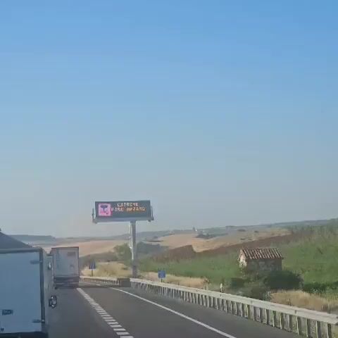 A digital sign above a highway displays "EXTREME FIRE HAZARD" in English and "RIESGO EXTRENO DE FUEGO" in Spanish, with a graphic of a burning tree. The sign is mounted on a pole and is visible to traffic on a sunny day with a clear blue sky. The surrounding landscape consists of dry, rolling hills and green vegetation. Several trucks are visible on the highway, and a small, rustic building is situated on the right side of the road.