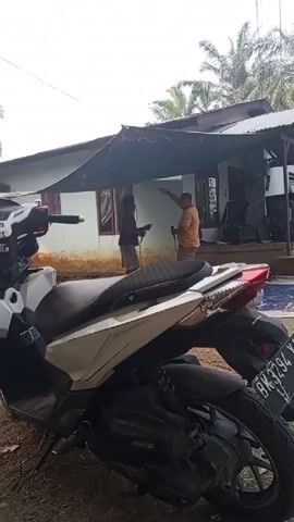 The video opens with a close-up of a white and beige motorcycle parked on a dirt surface. The background reveals a modest house with a tin roof and a tarpaulin extending over an outdoor area. Two men are visible under the tarpaulin. The man on the left is wearing a dark shirt and pants and is holding a plate. The man on the right is wearing an orange shirt and khaki pants and is holding a long pole or stick. He appears to be gesturing or pointing towards something off-camera. In the background, a third person, possibly a child, is sitting down. Palm trees are visible in the distance. The overall scene suggests an outdoor gathering or activity in a rural or semi-rural setting.