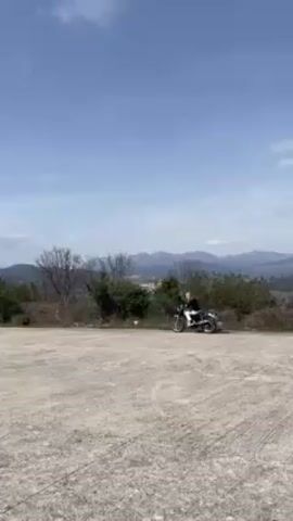 A woman is riding a motorcycle on a gravelly parking lot on top of a mountain. The woman has blonde hair and is wearing a black shirt and white pants. The motorcycle is black and silver. The background features mountains and trees. The sky is blue with a few clouds. The camera follows the woman riding the motorcycle slowly across the parking lot.