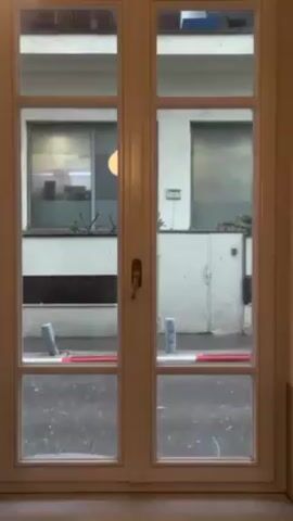 The video shows a view through a closed, white-framed window or glass door. The outside scene depicts a wet street, likely after rain, with reflections of the surrounding buildings visible on the pavement. A white car is briefly seen parked outside, then drives away. The buildings across the street are painted white, with windows, ledges, and some greenery. There are also some metallic gray bollards lining the edge of the street.