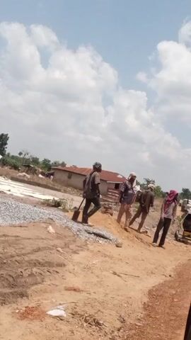 The video shows a group of four men walking along a dirt road in what appears to be a rural area. The men are wearing casual clothing, and some have head coverings. One man is carrying a shovel. Beside the road there's a pile of gravel and some construction materials. In the background, there are simple buildings and trees, under a sky with scattered clouds.
