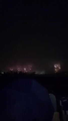 A nighttime video shows a crowd watching a fireworks display. In the foreground, the top of a dark blue umbrella is visible. Behind the umbrella, two people are seated, facing away from the camera, looking towards the fireworks. The person on the left is wearing a light blue shirt, and the person on the right is wearing a blue and white striped shirt with the word "LIONS" on the back. The sky is dark, with intermittent flashes of fireworks illuminating the scene with bright white, pink, and orange light. Smoke from the fireworks drifts across the sky. The display appears to be spread out horizontally across the horizon, with bursts of light visible in the distance above a dark line of trees or buildings.