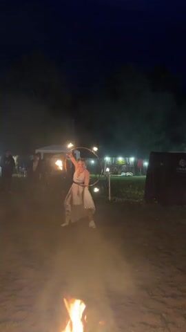 A woman in a white dress with gold trim and white boots is performing with a flaming hula hoop in a sandy, outdoor area at night. She has her hair up and is wearing glasses. The hula hoop has flames at multiple points around its circumference. There are other people in the background, some standing and some blurred. Lights are visible in the background, strung between structures that appear to be tents or booths. There are also fires on the ground in the foreground, with flames visible. The overall atmosphere appears to be an outdoor event or festival.