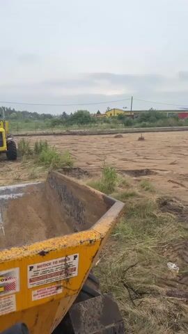 A first-person perspective video taken from the cab of a yellow dumper truck. The truck is on a dirt construction site with a cloudy sky. In the distance, two other vehicles are visible: a white truck and a yellow loader. The loader appears to be operated by a person wearing a safety vest. The dumper truck is filled with dirt and has various warning labels on its side. The scene suggests ongoing construction or earthmoving activities.